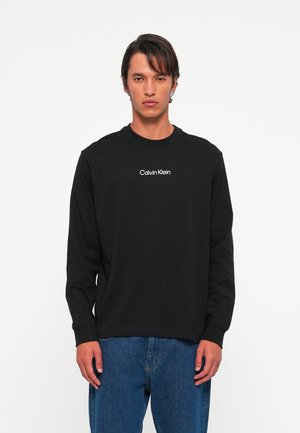 Man wearing black long-sleeve Calvin Klein shirt and blue jeans, standing against a plain white background.