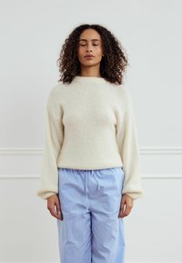 Kreminės spalvos megztinis su laisvu siluetu ir kamuolio formos rankovėmis, derinamas su mėlynomis dryžuotomis kelnėmis su raišteliu. Aiškiai jaučiamas minkštas tekstūros pojūtis.