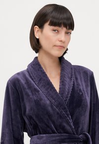 Lila, plysch badrock med en texturerad krage, bälte i midjan och långa ärmar. Mjuk tyg med subtil lyster, som har en liten logotyp.
