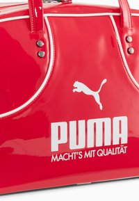 Punainen lakattu nahkalaukku valkoisilla yksityiskohdilla, jossa on lyhyet kahvat, metalliset niitit sekä rohkea musta Puma-logo ja teksti.