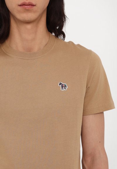 Beige bavlněné tričko s kulatým výstřihem, na levé straně hrudi s barevným vyšitým logem zebry. Krátké rukávy a hladká textura.