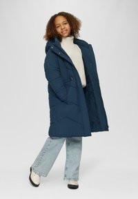 Navyblauer Daunenmantel mit Kapuze, mit einem gesteppten Design, Fronttaschen und einem weichen Futter, kombiniert mit hellblauen Schlagjeans und Schuhen.