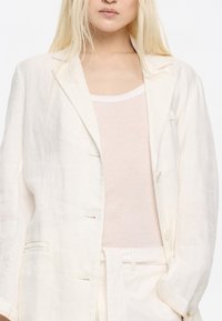 Ľanový blejzer v off-white farbe s priliehavým strihom, uzatváraním na jeden gombík a jemnými detailami. Nosený cez svetlý top bez rukávov.