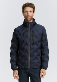 Man med marinblå vadderad jacka med hög krage, fram- och ärmfickor med dragkedja, står mot en enkel ljus bakgrund.