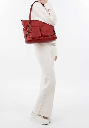Bolso tote de cuero rojo con dos bolsillos frontales y detalle de borla, combinado con pantalones ajustados de color crema y zapatillas blancas.