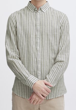 Homme portant une chemise à manches longues à boutons avec des rayures verticales beige et blanches, les mains jointes devant, un pantalon beige visible sous la chemise.