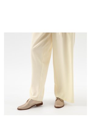 Beige perforierte Leder-Schnürschuhe, getragen mit locker sitzenden cremefarbenen Hosen vor einem einfarbigen Hintergrund.