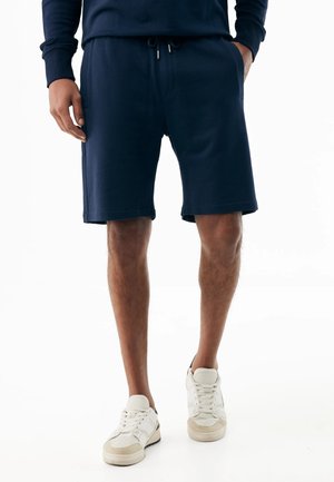 Mann trägt marineblaue Shorts mit Kordelzug, ein passendes Oberteil und weiße Turnschuhe, steht mit teilweise in die Taschen gesteckten Händen.
