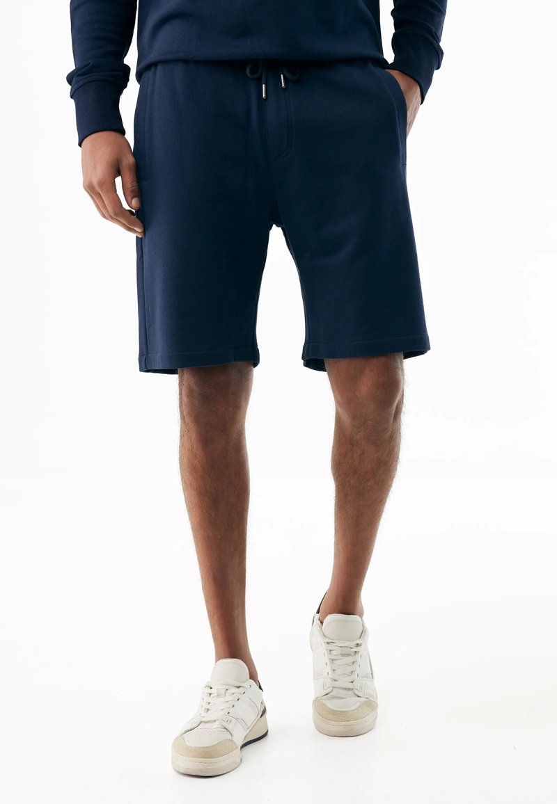 Man draagt marineblauwe shorts met trekkoorden, een bijpassend shirt en witte sneakers, staand met handen gedeeltelijk in de zakken.
