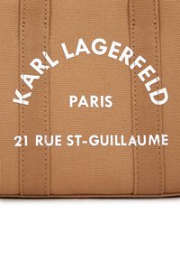 Tan tekstiltaske med hvidt printet tekst: "KARL LAGERFELD PARIS 21 RUE ST-GUILLAUME." Struktureret design med lodrette sømdetaljer.
