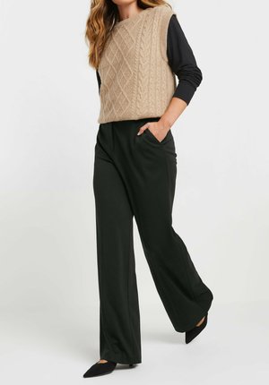 Beige Strickweste mit Zopfmuster, über einem schwarzen Langarmshirt getragen, kombiniert mit schwarzen, weit geschnittenen Hosen und spitzen schwarzen Ballerinas.