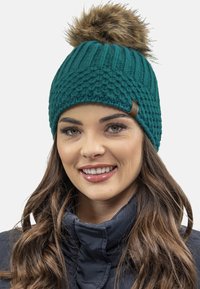 Bonnet tricoté en teal avec un motif texturé et un pompon en fausse fourrure marron sur le dessus. Présente une petite étiquette en cuir sur le côté.