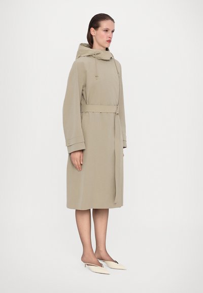 Beżowy płaszcz do kolan z kapturem, na sznurku i z paskiem w talii. Posiada szerokie rękawy i gładką fakturę. Para spiczastych beżowych butów.
