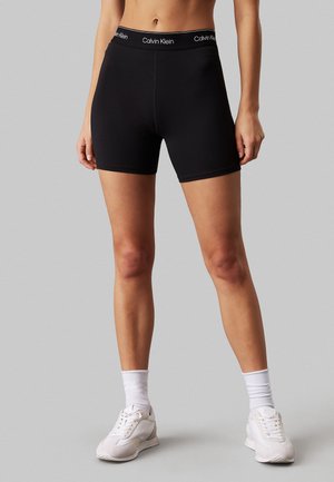 Pantaloncini elasticizzati neri con vita firmata Calvin Klein, design aderente, lunghezza a metà coscia e texture liscia. Indossati con sneakers bianche.