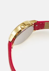 Goldfarbene Uhr mit rotem Lederarmband. Das Gehäuse ist rund mit einer glatten Oberfläche, verfügt über eine seitliche Krone und einen silbernen Rückdeckel.