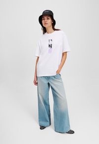 T-shirt bianco in cotone con stampa grafica, abbinato a jeans a gamba larga in denim chiaro. Un cappello a secchiello nero e scarpe a pois completano il look.