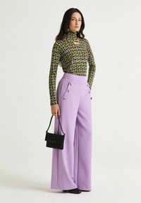 Top a manica lunga aderente con motivo geometrico verde e viola, abbinato a pantaloni larghi color lavanda con doppi bottoni, portando una borsa nera.