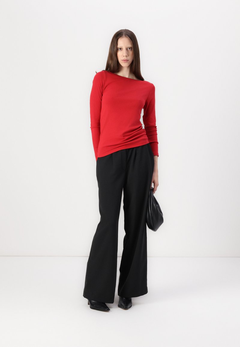 Haut rouge à manches longues avec un décolleté large, associé à un pantalon large noir. Le mannequin tient un sac à main noir et porte des talons aiguille noirs.