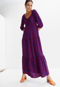 Abito maxi floreale blu e rosso con scollo a barca, arricciatura elastica sul busto, maniche a tre quarti e gonna a strati. Abbinato a tacchi arancioni.