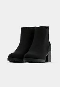 Un par de botas negras hasta el tobillo con tacones tipo bloque y paneles laterales elásticos sobre un fondo blanco liso.