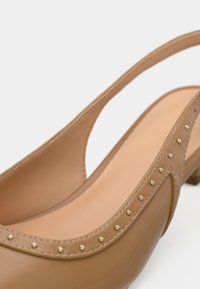 Μπεζ δερμάτινο γυναικείο παπούτσι slingback με στρογγυλεμένη μύτη, διακοσμητικά χρυσά καρφιά κατά μήκος της άκρης και χαμηλό τακούνι. Λείο, γυαλιστερό φινίρισμα με κοψίματα στο σχέδιο.