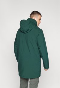 Grønn vanntett parkas med hette, med rett snitt, lange ermer og en glatt tekstur. Minimalistisk design uten synlige logoer.