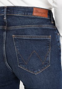 Tmavě modré džíny z denimu s hladkou texturou, které mají koženou nášivku a výrazné šikmé švy zadních kapes.