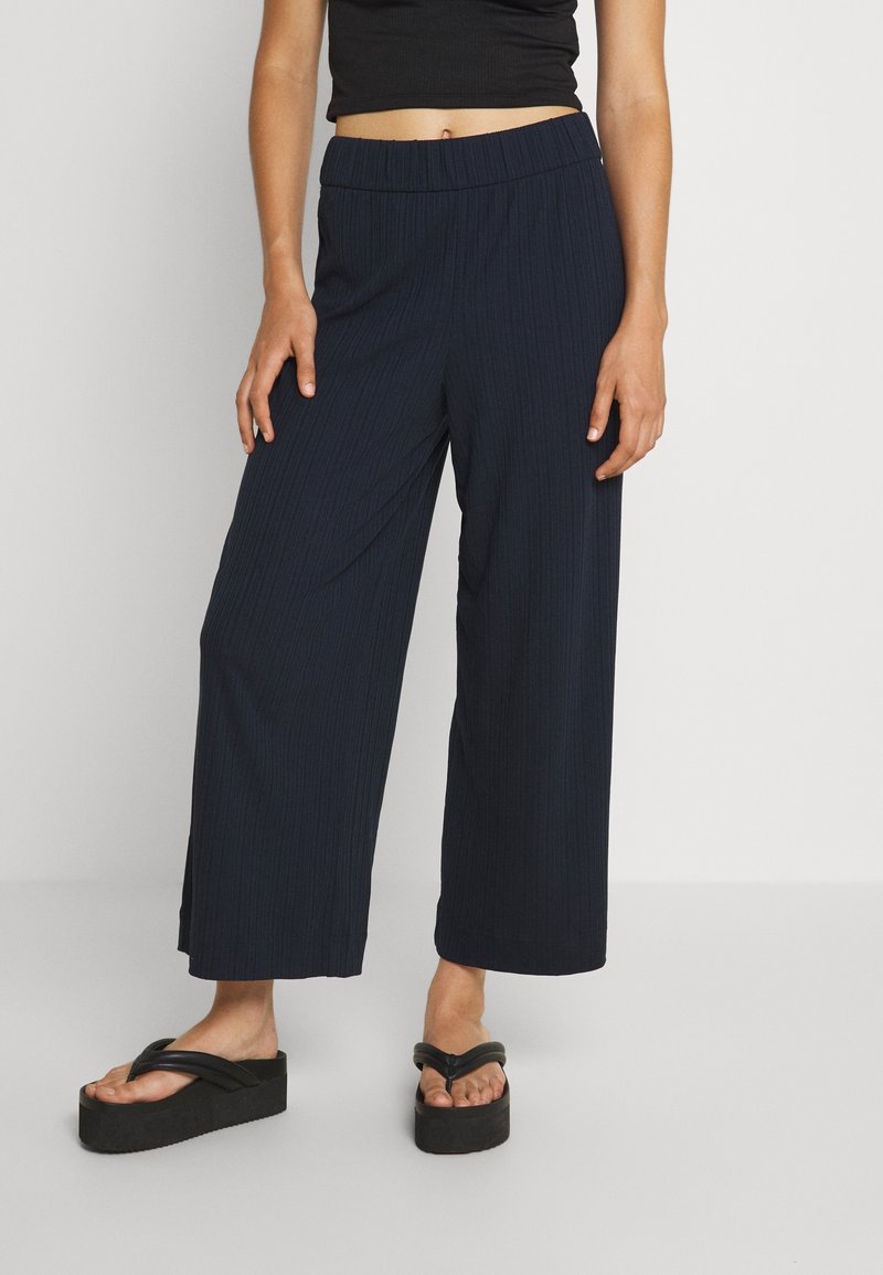 Femme portant un pantalon noir court à jambes larges, un top court noir et des tongs compensées noires, debout devant un fond gris clair.