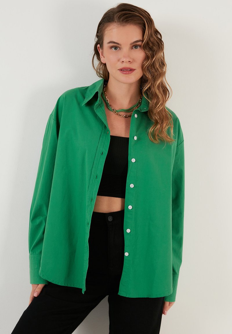 Green, button-up shirt made of a smooth fabric. Features long sleeves, a collar, and is worn open over a black cropped top.