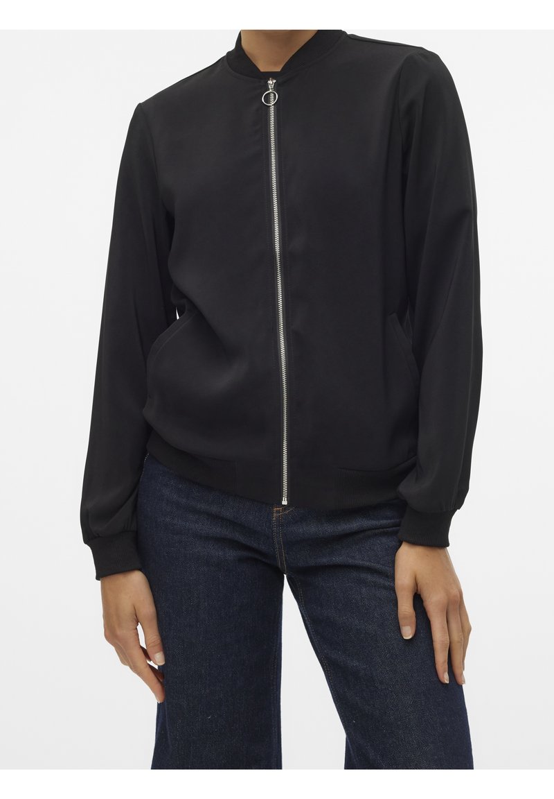 Personne portant une veste bomber noire zippée avec des poignets côtelés et un jean bleu foncé, debout devant un fond uni.