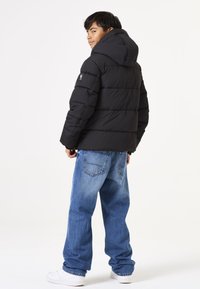 Chaqueta acolchada negra con capucha, con un patrón de acolchado vertical y un logo en la manga. Combinada con jeans azules sueltos y zapatillas blancas.