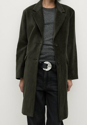 Persoon draagt een lange donkergroene jas, een grijze trui, zwarte jeans en een zwarte riem met een grote zilveren ronde gesp.