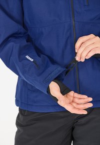 Die blaue wasserdichte Jacke verfügt über einen Klettverschluss an den Ärmeln und ein elastisches Innenfutter an den Handgelenken. Schwarze Akzente ergänzen das funktionale Design.