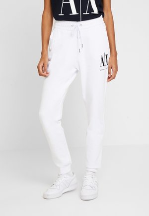 Osoba nosząca białe spodnie dresowe ze sznurkiem z czarnym logo "AX Armani Exchange" na udzie oraz białe trampki na jasnym tle.