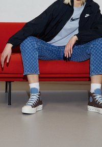 Bruna och blå högstøvlar med vita sulor, rutiga blå jeans, ljusblå t-shirt och svart vindjacka på en röd soffa.