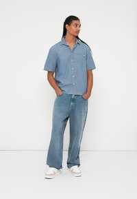 Cămașă din denim albastru cu mânecă scurtă și buzunar, purtată cu blugi ușor largi de un albastru deschis și adidași albi, pe un fundal simplu.