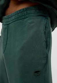 Groene joggingbroek van textuurstof met een losse pasvorm. Bevat een zijzak en een versleten gat voor extra detail.