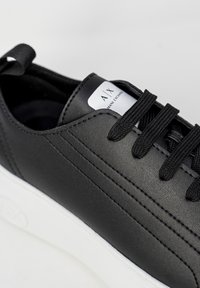 Detailní záběr na černou koženou tenisku s černými tkaničkami a bílou podrážkou, na bílém jazyku je viditelné logo Armani Exchange.