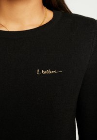 Svartstickad tröja med rund halsringning och guldstickad text som står "jag tror..." på vänster sida.