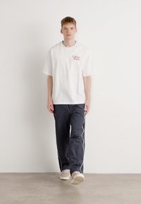 Witte T-shirt met korte mouwen en rood logo op de borst, gecombineerd met donkergrijze joggingbroek met witte bies en crèmekleurige sneakers.