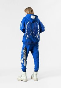 Sudadera azul con diseños de alas en la parte posterior, combinada con pantalones de chándal azules a juego que presentan una etiqueta gráfica en la pierna. Botines blancos.