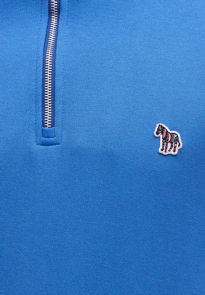 Μπλε πλεκτό ύφασμα με ένα μερικώς κλειστό ασημί φερμουάρ και ένα μικρό πολύχρωμο ριγέ μπαλώμα ζέβρας στη δεξιά πλευρά.