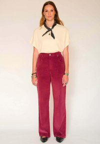 Pantalons larges en velours côtelé rouge avec poches avant, associés à un haut crème à manches courtes et une écharpe noire. Des chaussures noires à clous complètent le look.