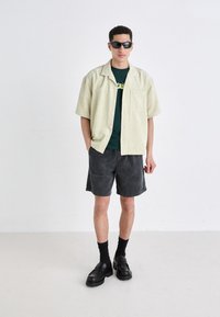 Lichtgroen shirt met korte mouwen, donkergroen t-shirt eronder, grijze shorts, zwarte chunky schoenen en een zonnebril, tegen een effen achtergrond.