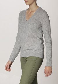 Pull en laine gris à col en V avec des poignets et un ourlet côtelés. Texture douce. Associé à un pantalon vert ajusté, montrant une tenue décontractée.