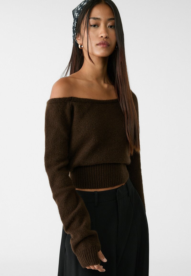 Brauner, schulterfreier Strickpullover mit geripptem Saum und langen Ärmeln; weicher Stoff und figurbetonter Schnitt. Kombiniert mit hochgeschnittenen schwarzen Hosen.