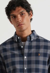 Chemise à carreaux bleu et gris avec col, dotée de boutons blancs et d'un accent de logo orange sur la poitrine. En matériau coton.