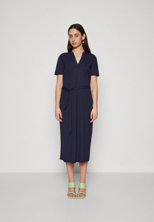 Vestido azul marino hasta la rodilla en tejido suave con escote en V, mangas cortas y cintura atada. Combinado con sandalias verdes gruesas.