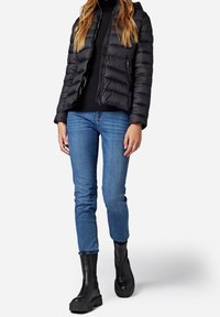 Černá péřová bunda s horizontálním prošíváním, zapínáním na zip a kapucí, kombinovaná s modrými slim jeans a černými kotníkovými botami s robustními podrážkami.