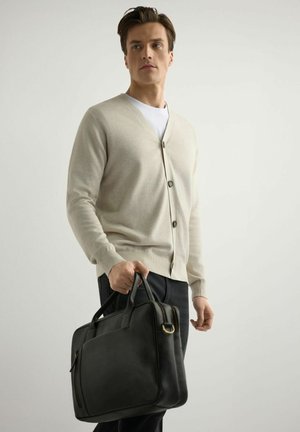 Młody mężczyzna w beżowym kardiganie i białej koszuli trzyma czarną skórzaną aktówkę na jasnym tle.